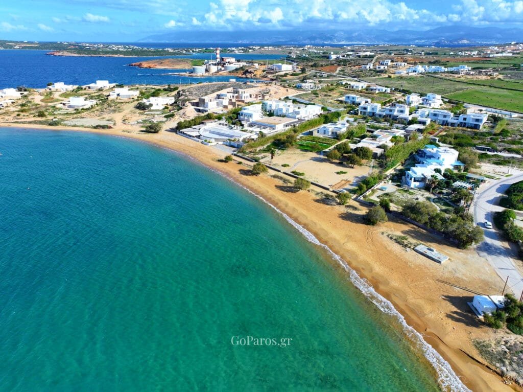 Agioi Anargyroi Beach, Paros, aerial view of the sandy shoreline with white villas, turquoise water, and an industrial chimney on the distant headland