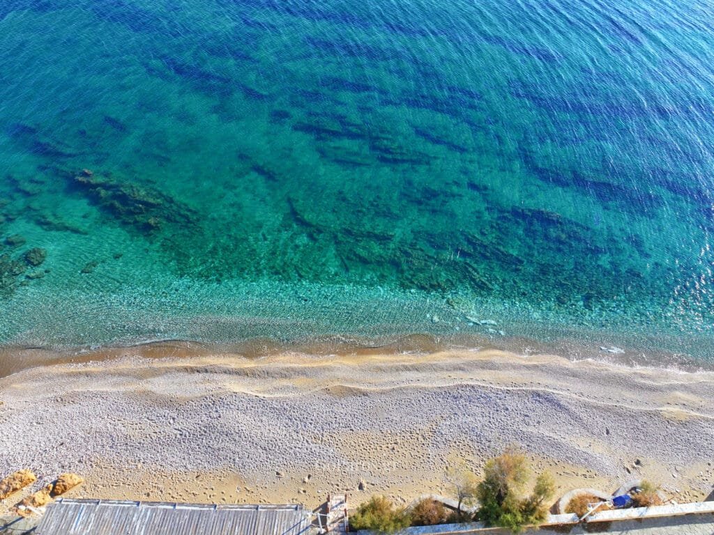 Top-down view of clear green-blue water meeting a sandy, pebbly shoreline at Drios Beach, Paros.