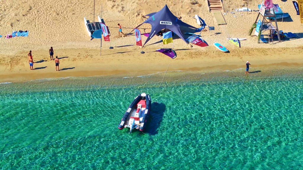 Drone shot of a RIB boat in shallow clear water at Golden Beach in Paros, in front of a watersports tent and people on the sand.