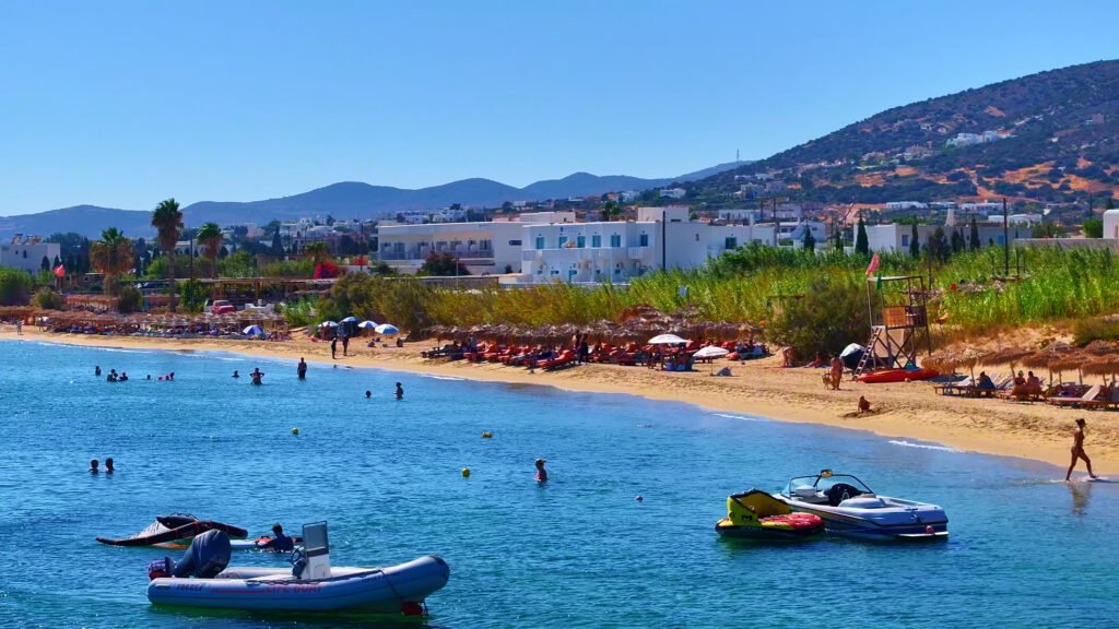 Golden Beach in Paros with swimmers in the water, small boats anchored near the shore and hotels and palm trees behind the sand.