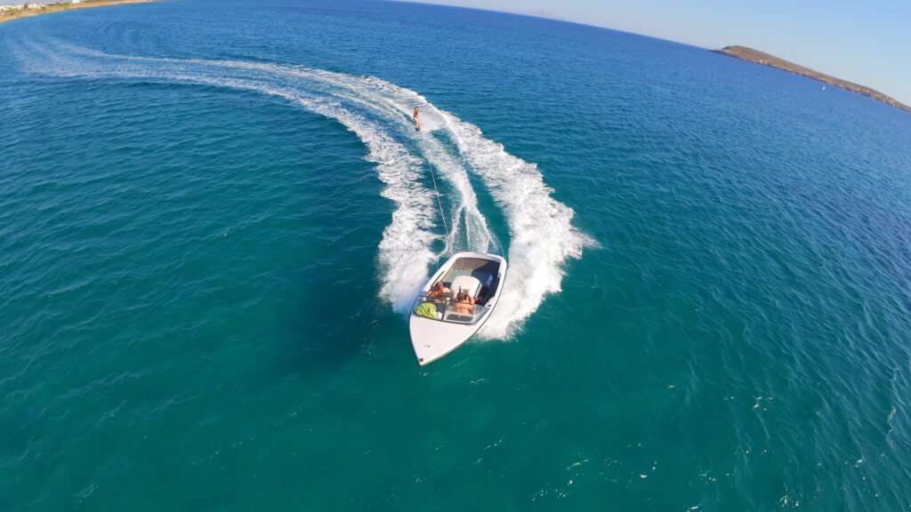 Drone view of a speedboat towing a waterskier off Golden Beach in Paros, leaving a curved white wake on the blue sea.