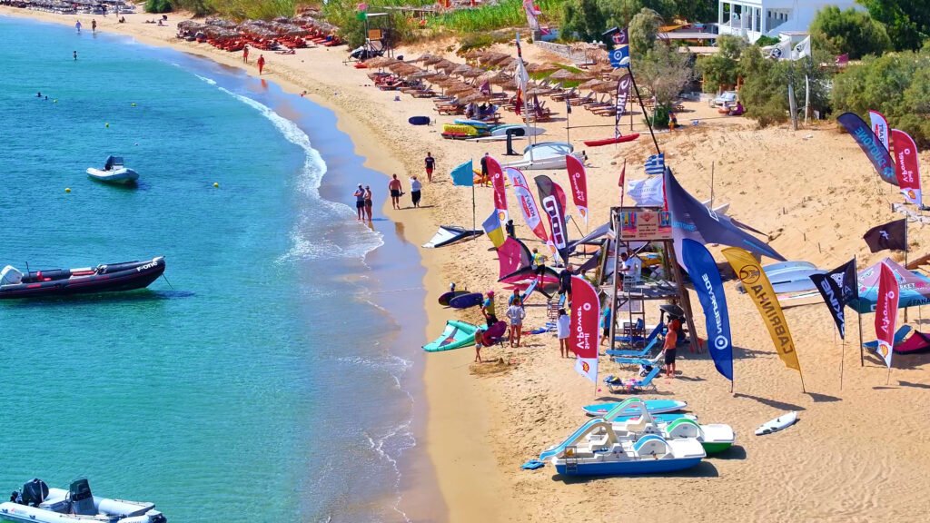 Golden Beach watersports center in Paros, with boards, kayaks and colorful flags set up on the sand next to the sea.