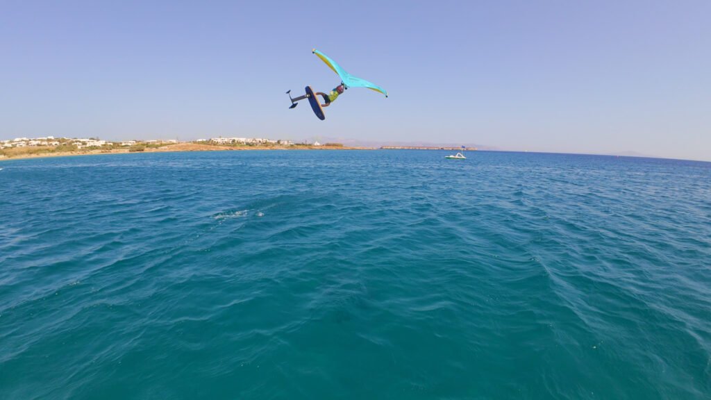 Wingfoiler catching air above the water off Golden Beach in Paros, with the coastline and low hills in the background.