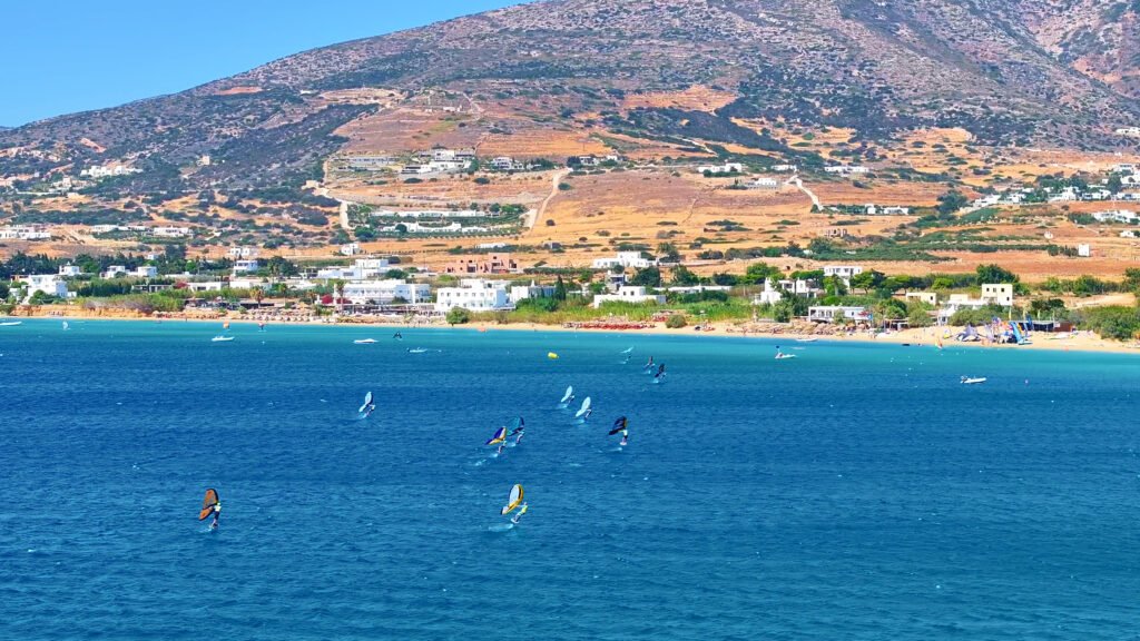 View of Golden Beach in Paros with multiple wink foilers crossing the bay and white houses and terraced hills behind the shoreline.