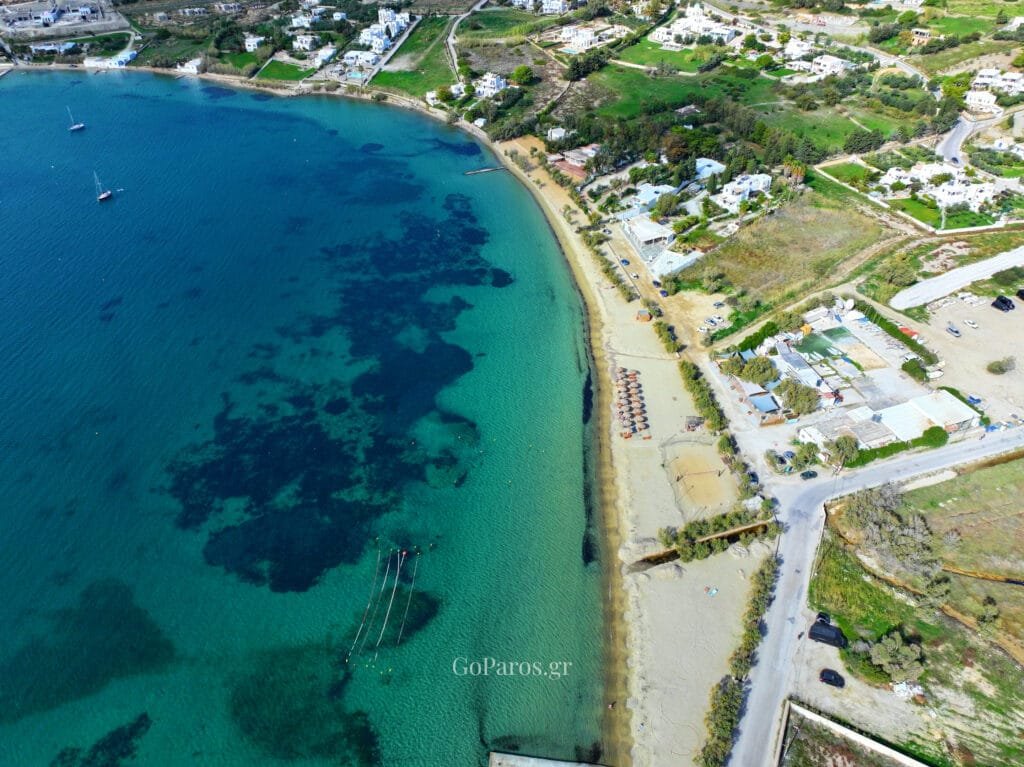 Livadia Beach, Paros, aerial view of the long sandy shoreline with beach umbrellas, a beach court, and clear water with dark seabed patches