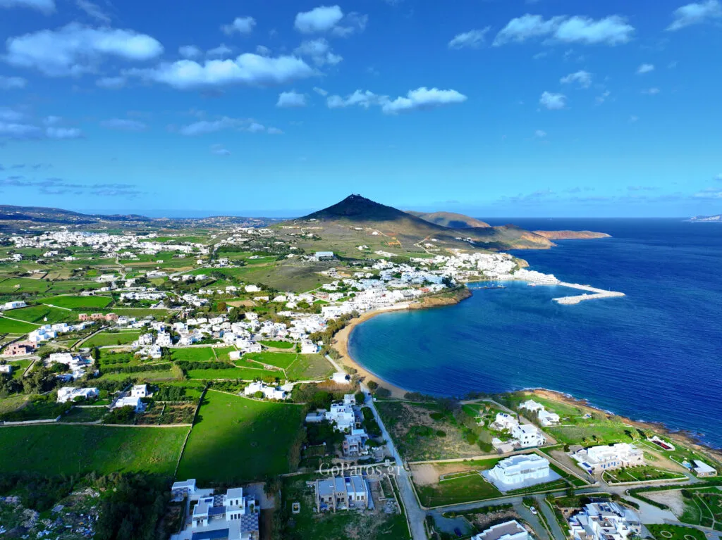 Aerial view of a crescent-shaped bay with a sandy shoreline, whitewashed buildings, and deep blue sea on Paros, Greece.