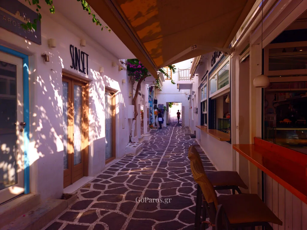Naoussa, Paros, narrow shopping alley with a cafe awning, window counter, and chairs on stone paving