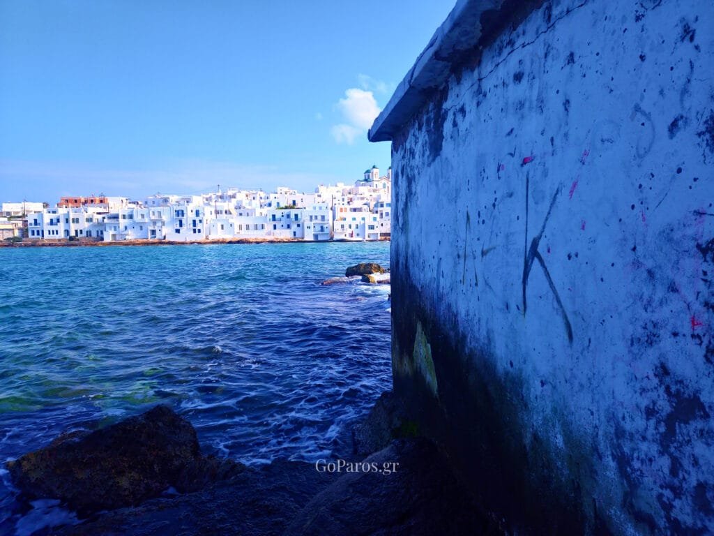 Naoussa, Paros, rocky shoreline and sea wall with white buildings across the water under a blue sky