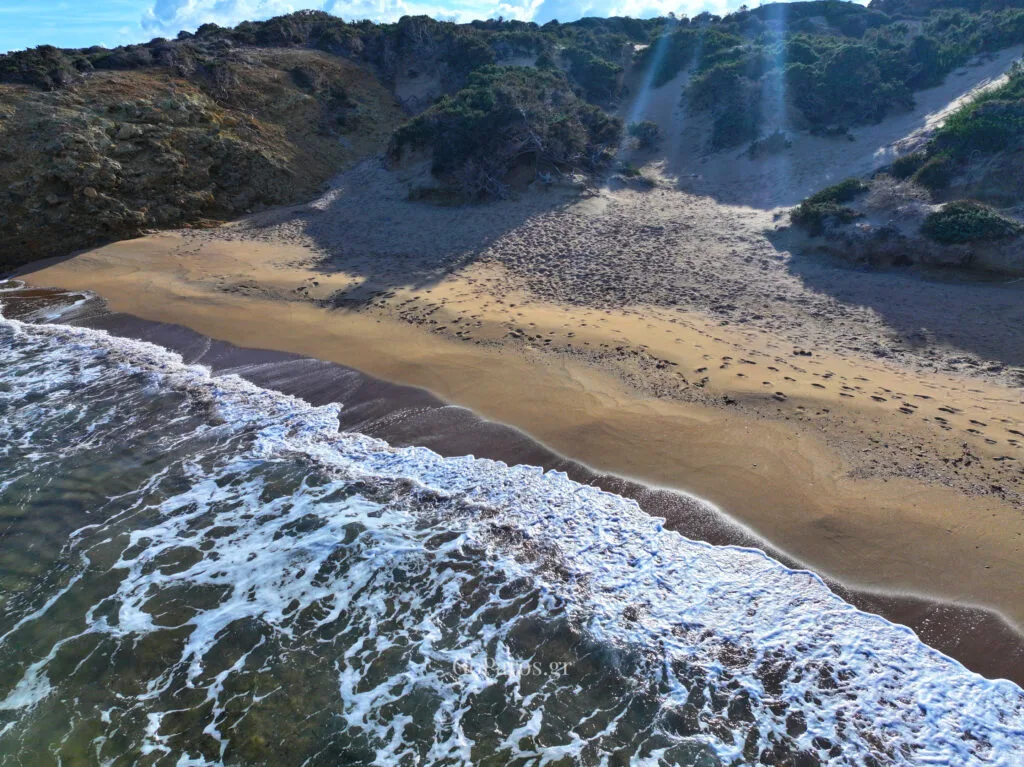 Palio Karnagio Beach, Paros, sandy dune hollow with shrubs and footprints above the surf, sun rays streaking across the scene