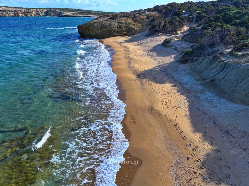 Palio Karnagio Beach, Paros, oblique view along the shoreline with small waves, footprints in sand, and rocky headland