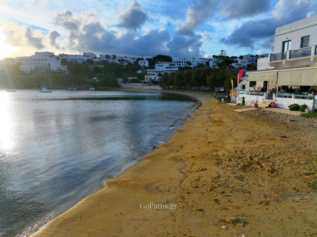 Piso Livadi Beach, Paros at sunset, sandy shoreline and calm water with waterfront buildings.
