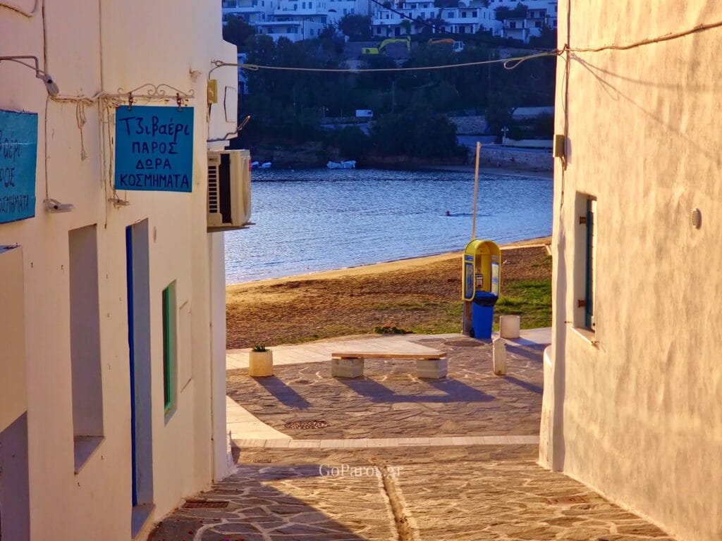 Piso Livadi, Paros, view down a narrow lane toward the beach with a yellow phone booth and benches by the shoreline.