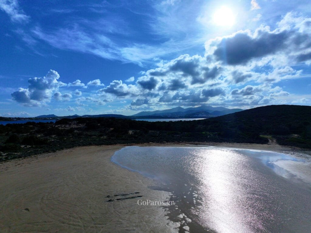 Platia Ammos Beach, Paros, low aerial along a narrow sandy beach with footprints, small waves, and a rocky cliff