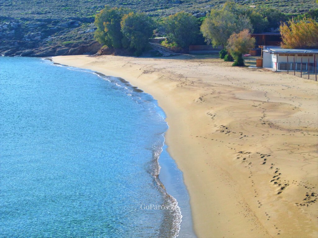 Curving sandy shoreline and calm sea at Punda Beach East, Paros, with beach facilities in the background.
