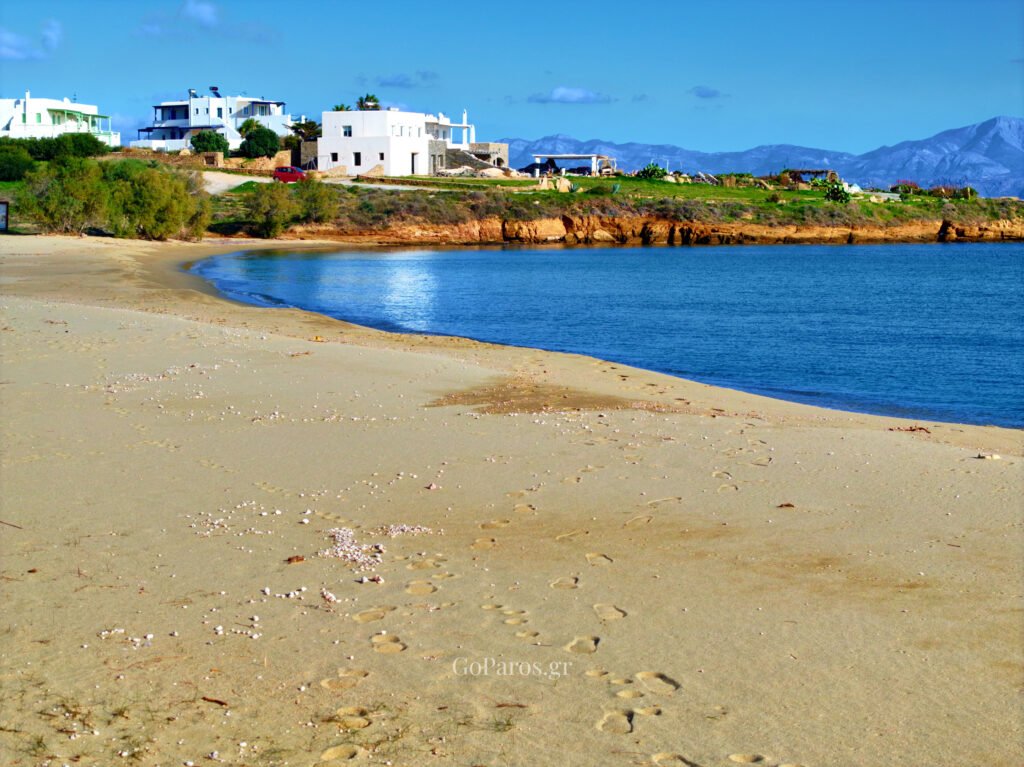 Quiet sandy shore at Punda Beach East, Paros, with white houses and coastline in the distance.