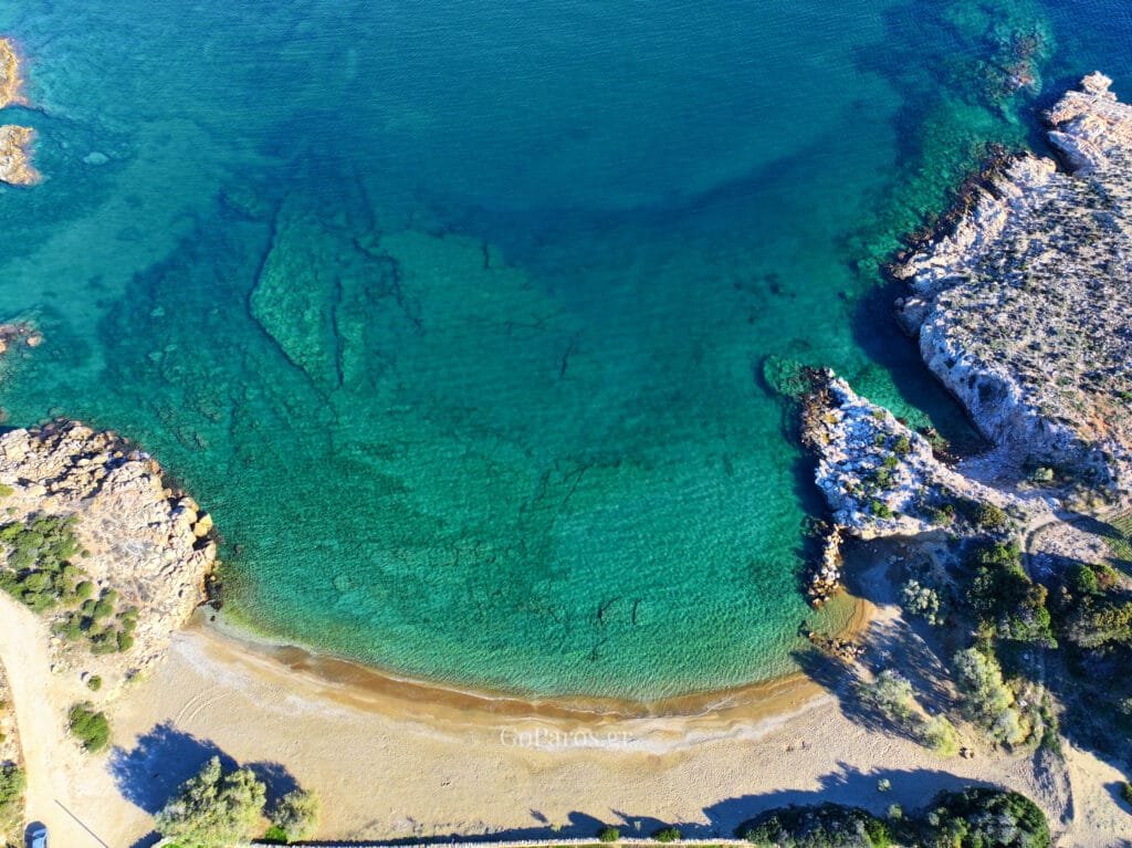 Aerial view of Tripiti Beach, Paros, showing a small sandy cove with crystal clear turquoise water and rocky edges.