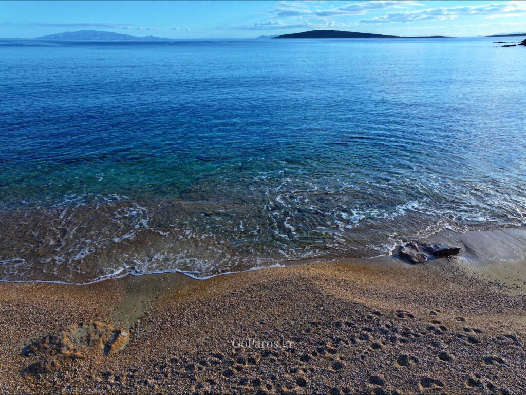 Makria Miti Beach, Paros, shoreline view with small waves on pebbles, footprints in the sand, and an islet offshore