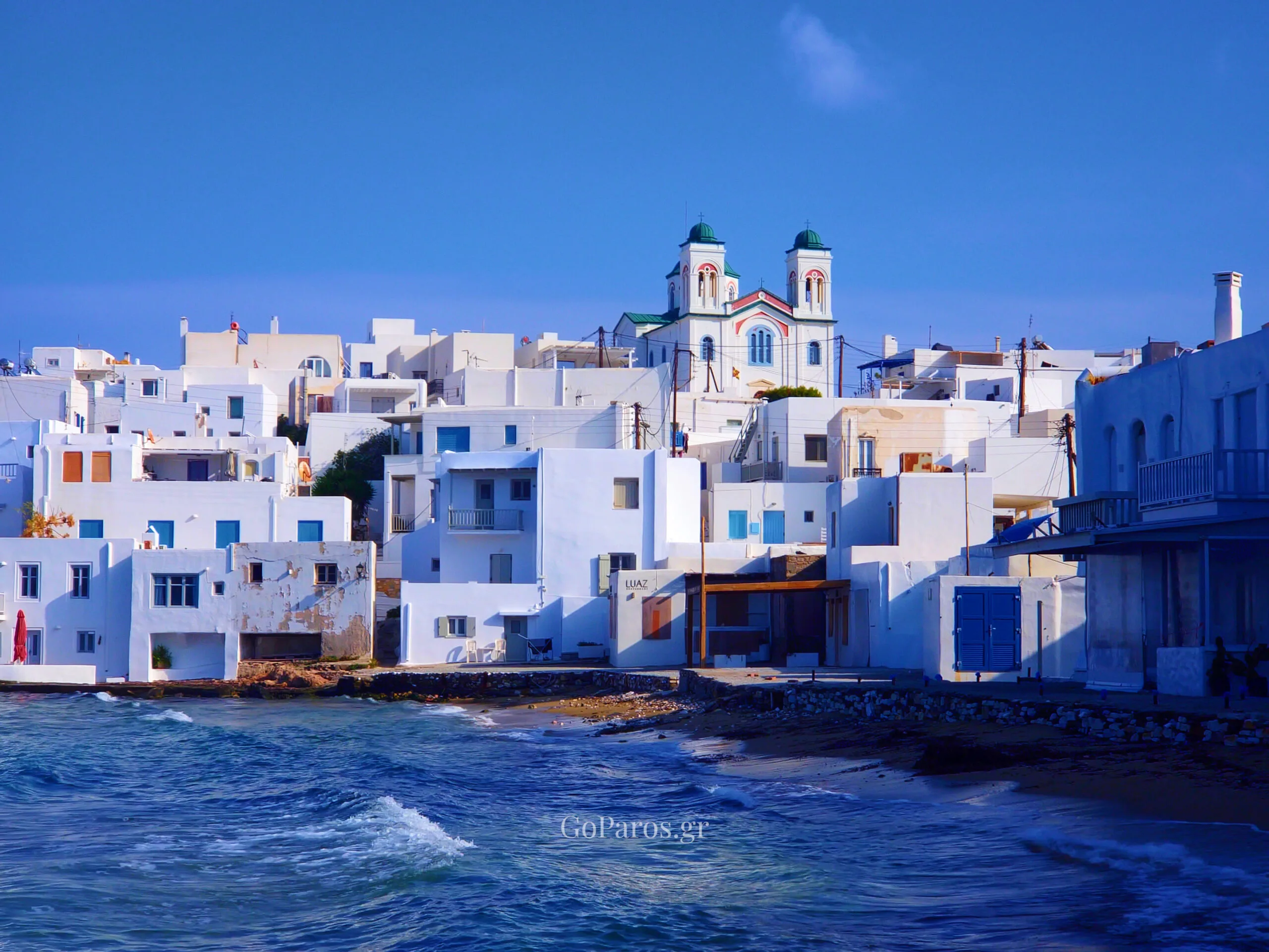 Naoussa, Paros seafront town view with white houses, a church, and waves along the shore